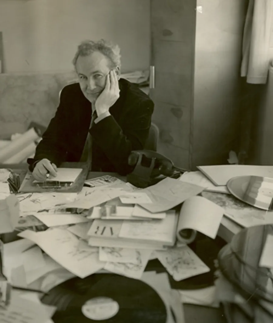 Bill Peet seated at his cluttered desk, resting his head on his hand among stacks of papers and drawings